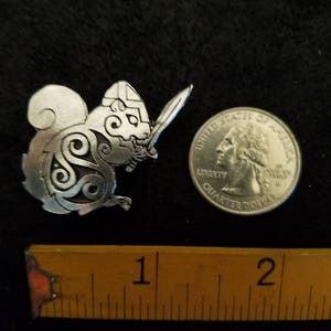 May include: A silver-colored metal squirrel-like figure with a sword, next to a quarter and a ruler. The squirrel has a helmet and Celtic knot designs. The ruler shows the figure is approximately 1.5 inches long.