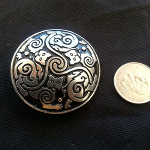 May include: A round, silver-toned metal brooch with a detailed design of stylized cats and swirling patterns. The brooch is next to a U.S. quarter coin for size comparison. The background is a dark fabric.