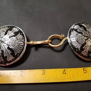 May include: A pair of silver and black metal clasps with a gold-toned connecting piece. The clasps feature a Celtic knot design with a leaf motif.