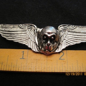 May include: Silver monkey head with wings pin. The monkey has a serious expression and is wearing a pair of wings. The pin is about 1 inch wide.