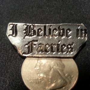 May include: A silver charm with the words "I Believe in Fairies" engraved in black lettering. The charm is about the size of a US quarter.