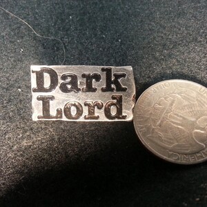 May include: A silver metal pin with the words "Dark Lord" in black lettering.
