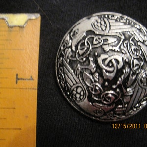 May include: A silver-toned metal brooch with a Celtic knot design. The brooch features intricate details and a raised design.
