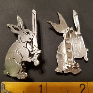May include: Silver-toned rabbit pin with a sword in its paw. The back of the pin has a safety clasp.