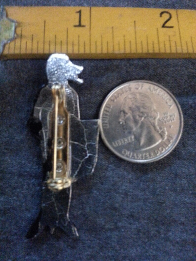 Puede incluir: Un broche negro y plateado con forma de mujer. El broche es aproximadamente del tama&ntilde;o de una moneda de 25 centavos.