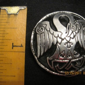 May include: A silver-toned metal pin with a black enamel background. The pin features a detailed design of a bird with outstretched wings, set against a Celtic knot pattern.