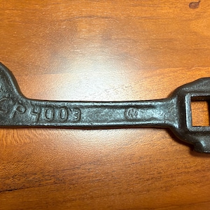 May include: A dark gray metal wrench featuring an open-ended head on one side and a square-shaped socket head on the other. The tool has the embossed text CP 4003 and a small circular logo stamped on the central shaft of the metal body.