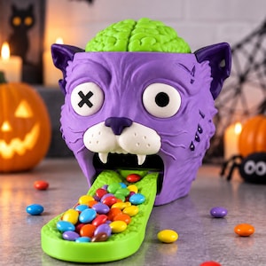 May include: A purple cat-shaped sweet dispenser with a bright green brain on top and a long green tongue extending from its mouth. The cat has one white eye with a black X and one solid black eye. Small colourful sweet pieces are on the tongue.