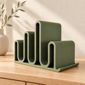 May include: A matte olive green desk organizer featuring a series of four vertical U-shaped slots of varying heights. The structure is made of a solid material with a smooth finish, designed to hold papers, mail, or documents in an upright position.