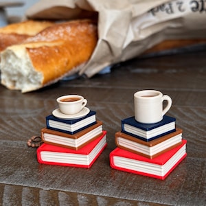 May include: Two miniature stacks of three books each, topped with small cups. The left stack has a wide, shallow cup on a saucer, while the right stack features a taller mug. The books are colored red, brown, and dark blue with white pages.