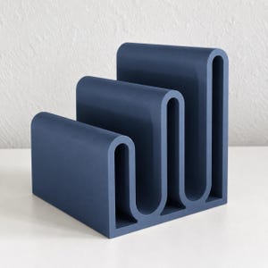 May include: A matte navy blue desk organizer featuring a stepped, three-tier design with continuous, rounded U-shaped dividers. The structure is composed of three distinct sections of varying heights, creating a functional tiered storage unit.