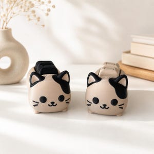 May include: Two cat-shaped tape dispensers made of beige material with black ears, eyes, whiskers, and mouth details. One dispenser features a black tape roll holder, while the other has a beige tape roll holder, both positioned on the cat's back.