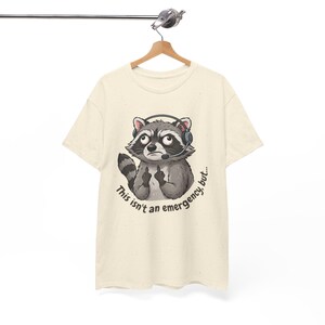 May include: A cream-coloured short-sleeve t-shirt featuring a graphic of a raccoon wearing a headset. The raccoon is making a rude gesture with both hands. Below the raccoon, the text reads: This isn't an emergency, but...