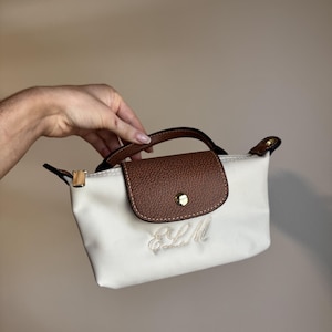 May include: A small off-white handbag featuring a brown textured flap with a gold-tone snap closure, a top handle, and the embroidered monogram letters ELM in a cursive script on the front panel of the bag, accented with gold-tone hardware.