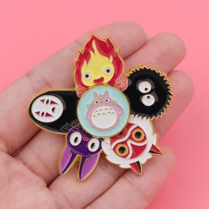 May include: A gold-toned metal enamel pin featuring a central light blue circle with a pink creature, surrounded by five distinct character designs including a red flame, a black mask, a black fuzzy sphere, a purple rabbit, and a white mask.