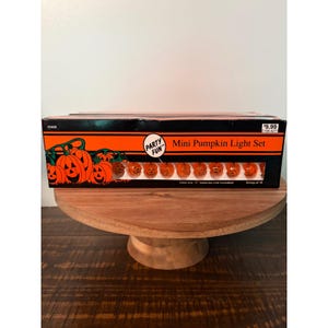 May include: A rectangular black and orange box labeled Party Fun Mini Pumpkin Light Set. The packaging features illustrations of jack-o'-lanterns and a clear window displaying a string of ten small orange pumpkin-shaped lights inside.