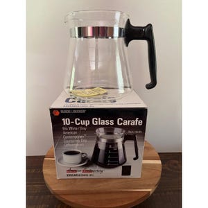 May include: A clear glass coffee carafe with a black plastic handle and a silver metal band around the top. The carafe sits on its original cardboard box, which features the text 10-Cup Glass Carafe, Black & Decker, and 3351AC(679419-01).