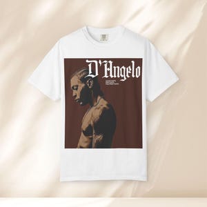 Può includere: Una t-shirt bianca a maniche corte con una grande grafica rettangolare sul petto. La grafica presenta uno sfondo marrone con un ritratto di D'Angelo e il testo D'Angelo, Good Days, Bad Days, Halfway Days in caratteri bianchi.