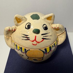 May include: A round, cream-colored ceramic cat figurine with a hand-painted design. The cat features a green spot on its head, black eyes, a green nose, and a red mouth. It has raised paws, a yellow collar, and blue and yellow decorative patterns.