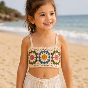 May include: A cream-colored crochet crop top featuring three prominent floral granny square motifs in shades of pink, olive green, yellow, and blue. The garment has thin shoulder straps and a scalloped edge along the top neckline.