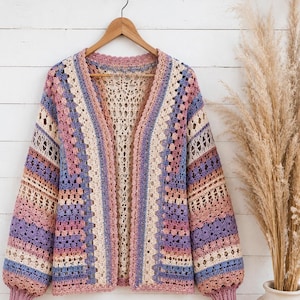 Può includere: Un cardigan all'uncinetto a maniche lunghe con un motivo a righe multicolore nei toni del rosa polvere, lavanda, blu e crema. Il capo presenta un'apertura frontale, uno scollo a V e polsini a coste, realizzato con punti pieni e traforati.