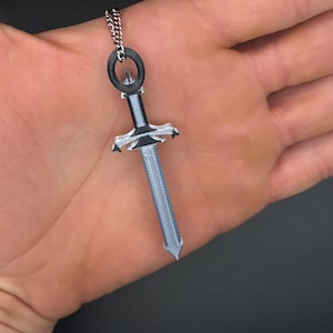 Può includere: Un ciondolo a forma di spada color argento appeso a una sottile catena di metallo. La spada presenta un'impugnatura e una guardia nere con una lama grigio chiaro testurizzata. Il design include un anello circolare in cima per la catena.