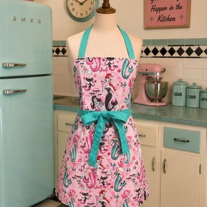 Peut inclure: Un tablier rose orné d'un motif répétitif de sirènes et de chats noirs, agrémenté de bretelles et d'un lien de taille en tissu bleu sarcelle uni. Le tissu présente diverses silhouettes de sirènes aux queues colorées, des poissons et des étoiles.