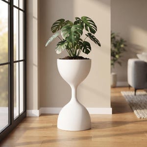 May include: A white hourglass-shaped floor planter with a smooth, matte finish. The planter features a wide, rounded top section holding a green Monstera plant with large, split leaves and a narrow, tapered middle section resting on a wide base.