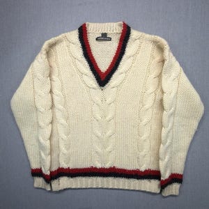 May include: A cream-colored cable knit sweater featuring a V-neckline with red and navy blue striped trim. The long-sleeved garment includes matching red and navy blue horizontal stripes along the ribbed hem and cuffs, with a visible brand label.