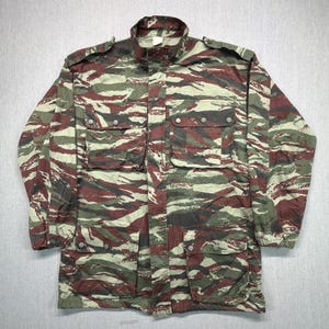May include: A long-sleeve jacket featuring a tiger stripe camouflage pattern in shades of dark red, olive green, and tan. The jacket includes a high collar, shoulder epaulets, and four large front pockets with button-down flaps for secure storage.