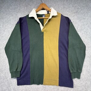 Może przedstawiać: Koszulka do rugby z długim rękawem o wzorze typu color-block w kolorach ciemnej zieleni, granatu i musztardowej żółci. Koszulka posiada biały kołnierzyk, zapięcie na guziki oraz metkę marki GAP przy dekolcie. Rękawy są ciemnozielone i granatowe.