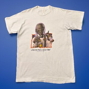 May include: A white short-sleeve t-shirt featuring a graphic of a creature in a tuxedo holding two beer bottles. The text below the graphic reads JOIN ME FOR A COLD ONE! The creature has long hair and a pale complexion, set against an orange border.