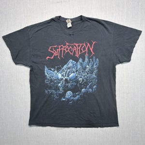 Pode incluir: Uma t-shirt preta de manga curta com uma impressão gráfica no peito. O design inclui a palavra "SUFFOCATION" em letras vermelhas estilizadas sobre uma ilustração detalhada em azul e branco de uma cena caótica com figuras e estruturas.