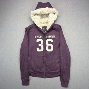 May include: A purple zip-up hoodie featuring a cream-coloured faux fur lining in the hood. The front displays the text ABERCROMBIE above the number 36 in large cream-coloured lettering. The garment includes long sleeves and a full-length front zipper.