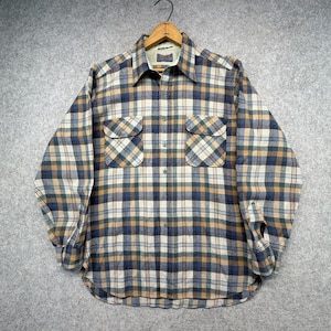 May include: A long-sleeve button-down shirt featuring a plaid pattern in shades of tan, navy blue, and white. The shirt includes a pointed collar, two chest pockets with buttoned flaps, and a front button closure. The fabric has a soft texture.