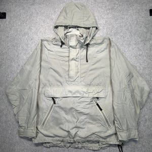 Könnte beinhalten: Eine hellbeige Kapuzen-Pulloverjacke mit einer großen horizontalen Fronttasche mit Klappe, zwei diagonalen Reißverschlusstaschen darunter, langen Ärmeln mit verstellbaren Bündchen sowie schwarzen Kordelzügen an Kapuze und Saum.