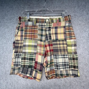 May include: Multi-colored plaid shorts featuring a patchwork design with various square panels in shades of red, yellow, green, blue, and cream. The shorts include a waistband with belt loops, a single front button closure, and a zipper fly.
