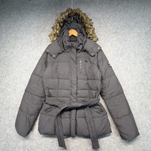 Puede incluir: Una chaqueta acolchada de color gris oscuro con capucha y ribete de piel sintética marrón, cierre de cremallera frontal, cinturón a juego en la cintura y un pequeño bolsillo con cremallera en el pecho. Mangas largas y diseño acolchado.