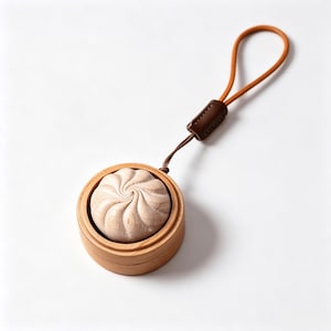 May include: A light-colored wooden charm shaped like a steamed bun, nestled inside a small, round, light-colored wooden container. The charm is attached to a thin, tan-colored leather cord with a dark brown leather connector loop for hanging.