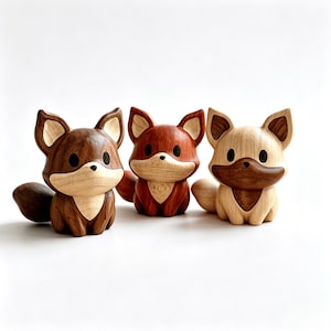 May include: Three carved wooden fox figurines in different wood tones. The left fox is dark brown, the centre fox is reddish-brown, and the right fox is light tan. Each fox features large black eyes, pointed ears, and a smooth, polished finish.