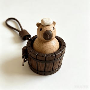 Könnte beinhalten: Eine hölzerne Capybara-Figur mit einem kleinen weißen, strukturierten Block auf dem Kopf, die in einem dunkelbraunen Holzfass sitzt. Das Fass verfügt über einen Metallring, der mit einer dunkelbraunen Lederschlaufe als Schlüsselanhänger verbunden ist.