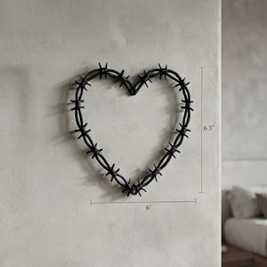 Op de afbeelding: Een hartvormige wanddecoratie gemaakt van zwart prikkeldraad. Het object is voorzien van een reeks scherpe metalen punten langs het draadframe. De aangegeven afmetingen op de afbeelding zijn 16,51 cm hoog en 182,88 cm breed.