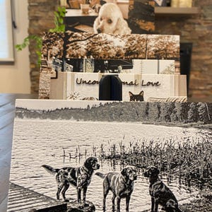 K&ouml;nnte beinhalten: Eine rechteckige Schieferplatte mit einer lasergravierten Schwarz-Wei&szlig;-Abbildung von drei Hunden an einem Seeufer neben einem Holzsteg. Die strukturierte Oberfl&auml;che zeigt detaillierte Silhouetten der Hunde vor einem Hintergrund aus Wasser und Schilf.
