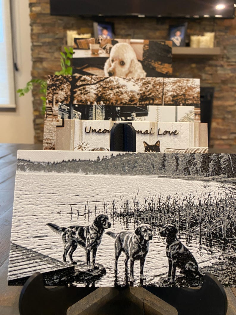 K&ouml;nnte beinhalten: Eine rechteckige Schieferplatte mit einer lasergravierten Schwarz-Wei&szlig;-Abbildung von drei Hunden an einem Seeufer neben einem Holzsteg. Die strukturierte Oberfl&auml;che zeigt detaillierte Silhouetten der Hunde vor einem Hintergrund aus Wasser und Schilf.