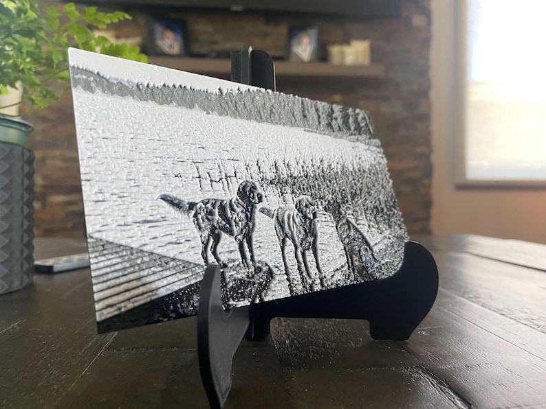 K&ouml;nnte beinhalten: Eine rechteckige Metallplatte mit einer schwarz-wei&szlig;en Gravur von drei Hunden, die auf einem Holzsteg an einem See stehen. Die Platte wird auf einem schwarzen Kunststoffst&auml;nder pr&auml;sentiert und zeigt eine detaillierte, strukturierte Oberfl&auml;che.