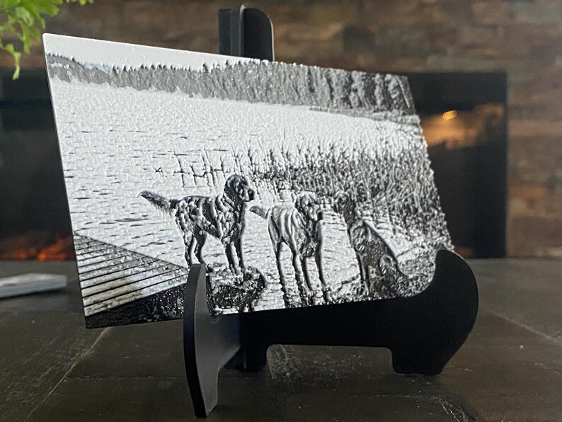K&ouml;nnte beinhalten: Eine rechteckige schwarz-wei&szlig;e Metallplatte mit einer Gravur von drei Hunden, die auf einem Holzsteg an einem See stehen. Die Platte wird auf einem schwarzen Kunststoffst&auml;nder pr&auml;sentiert und zeigt eine detaillierte Landschaft mit Wasser.