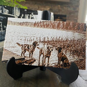 K&ouml;nnte beinhalten: Eine rechteckige Holzplatte mit einem sepiafarbenen, lasergravierten Bild von drei Hunden, die an einem Seeufer mit Schilf und B&auml;umen im Hintergrund stehen. Die Platte wird auf einem schwarzen Kunststoffst&auml;nder pr&auml;sentiert und zeigt Details.
