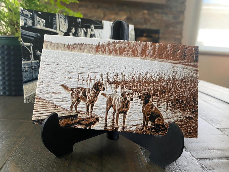 K&ouml;nnte beinhalten: Eine rechteckige Holzplatte mit einem sepiafarbenen, lasergravierten Bild von drei Hunden, die an einem Seeufer mit Schilf und B&auml;umen im Hintergrund stehen. Die Platte wird auf einem schwarzen Kunststoffst&auml;nder pr&auml;sentiert und zeigt Details.