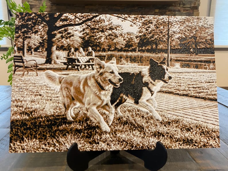 K&ouml;nnte beinhalten: Eine rechteckige Holzplatte mit einer lasergravierten, sepiafarbenen Abbildung von zwei Hunden, die &uuml;ber eine Wiese in einem Park rennen. Im Hintergrund sind ein Ziegelweg, B&auml;ume, eine Bank und Menschen an einem Picknicktisch an einem Gew&auml;sser zu sehen.