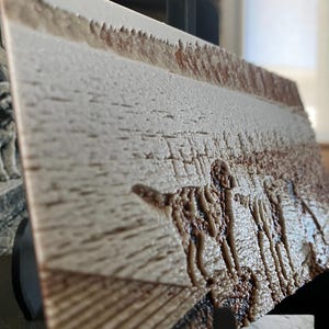 K&ouml;nnte beinhalten: Eine rechteckige Platte mit einem strukturierten Reliefbild eines Hundes, der auf einem Feld steht. Die Oberfl&auml;che hat eine cremefarbene und hellbraune Farbpalette mit einer rauen, taktilen Textur und steht auf einem schwarzen St&auml;nder.